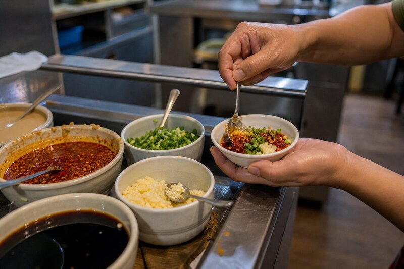 A hotpot condiment station with bowls of sesame paste, chili oil, minced garlic, spring onions, and soy sauce for building dipping sauces