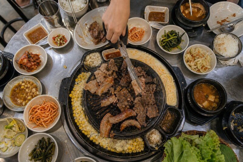 A Korean BBQ table mid-meal: marinated pork and beef grilling over a central dome grill, tongs and scissors in hand, ringed by small dishes of kimchi, corn cheese, and pickled sides