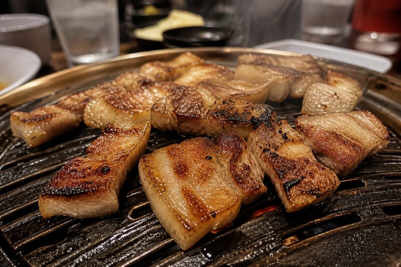 Thick slices of pork belly caramelizing on a Korean BBQ grill, edges crisping and glistening