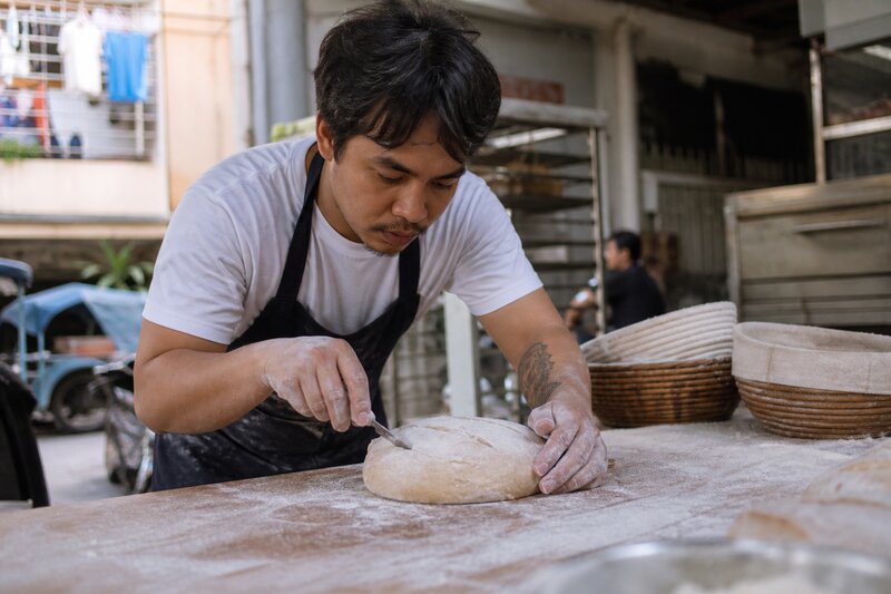 A baker shaping and scoring dough on a flour-dusted work surface in an open bakery kitchen